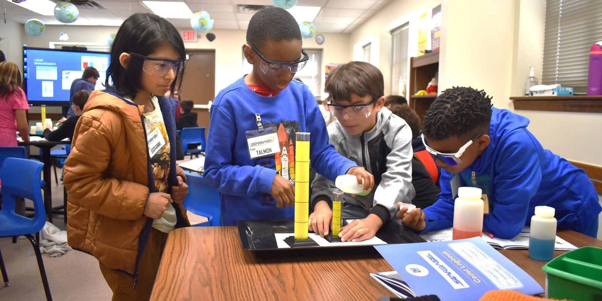 Students gathered around a table measuring liquid in graduated cylinders during a hands-on activity.