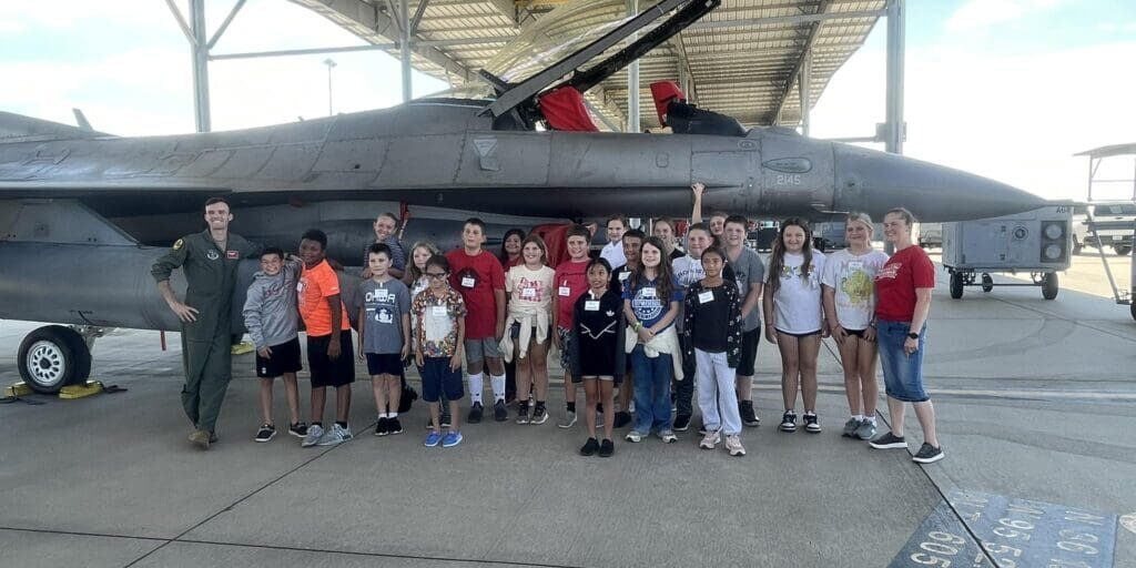 Group of children posing with an F-16 pilot in front of a parked fighter jet.